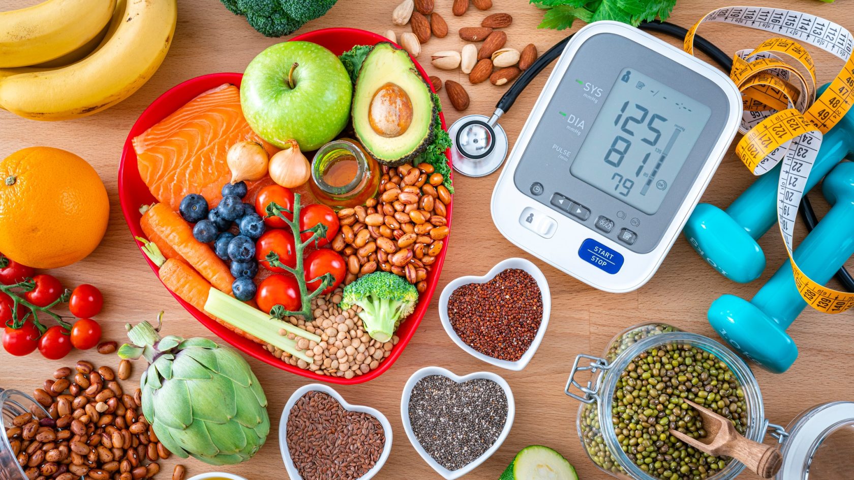 Heart care concept. Overhead view of a group of food that contains healthy nutrients for heart care, a blood pressure monitor, dummbells, tape measure and sthetoscope shot on wooden table. The composition includes extra virgin olive oil, legumes, avocado, berries, nuts and seeds, salmon, broccoli, carrot, soy beans, brown lentils, eggplent, spinach, oats, artichoke, apple, tomato, garlic among others. High resolution 42Mp studio digital capture taken with SONY A7rII and Zeiss Batis 40mm F2.0 CF lens